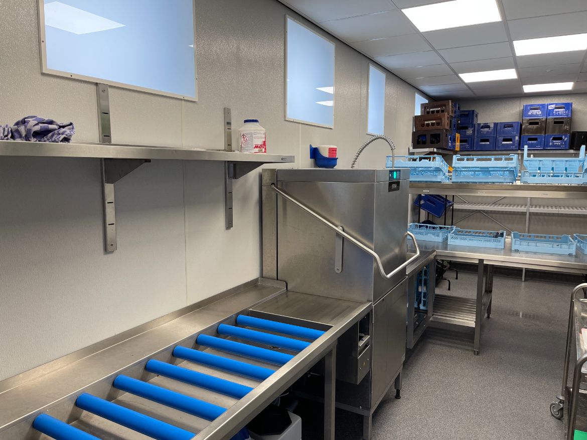 Zone de plonge et laverie d'une cuisine professionnelle, murs habillés en panneaux composites blancs derrière le convoyeur et la douchette